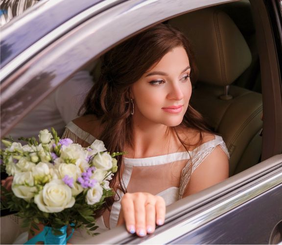 A bride in a white wedding dress holding a bouquet of white and purple flowers, sitting inside a limousine