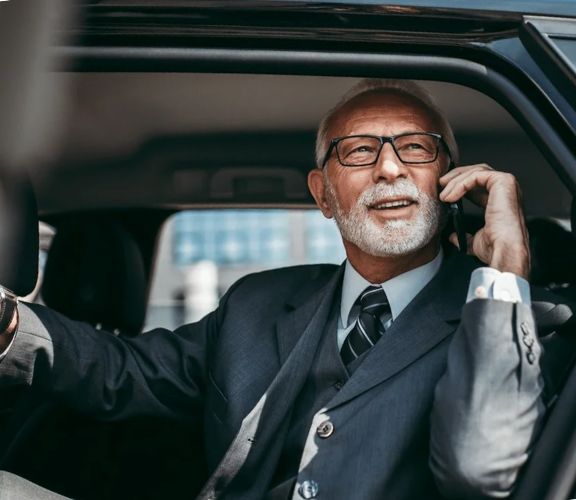 A businessman in a suit talking on a phone while sitting in a car with the door open, with a cityscape in the background.