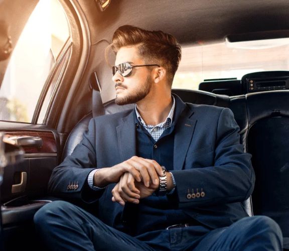 A man in a suit sitting inside a luxurious Limo, looking out the window with sunlight streaming in, hands clasped, wearing sunglasses and a watch.