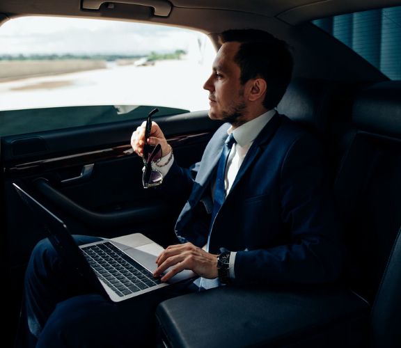 A businessman working on a laptop and holding sunglasses inside a luxury limo.