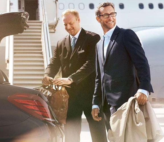 Chauffeur in a dark suit placing a traveler’s bag in the trunk during an airport transportation Bridgeport CT pickup, aircraft stairs behind them