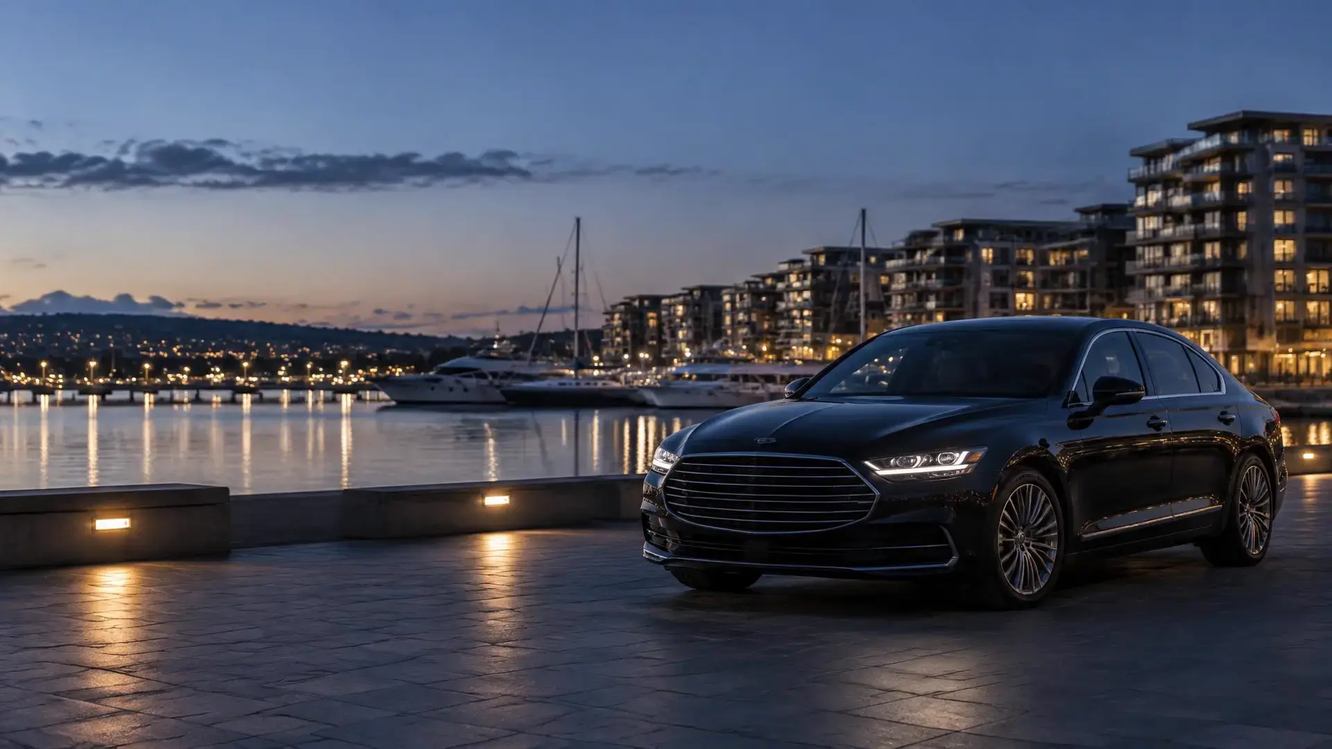 A black luxury sedan parked on a waterfront promenade at dusk, with moored boats and lit apartment buildings across the harbor