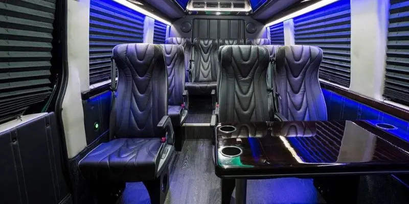 Inside a passenger van, quilted blue-gray leather seats, a black table with cup holders, window blinds and blue ambient lighting